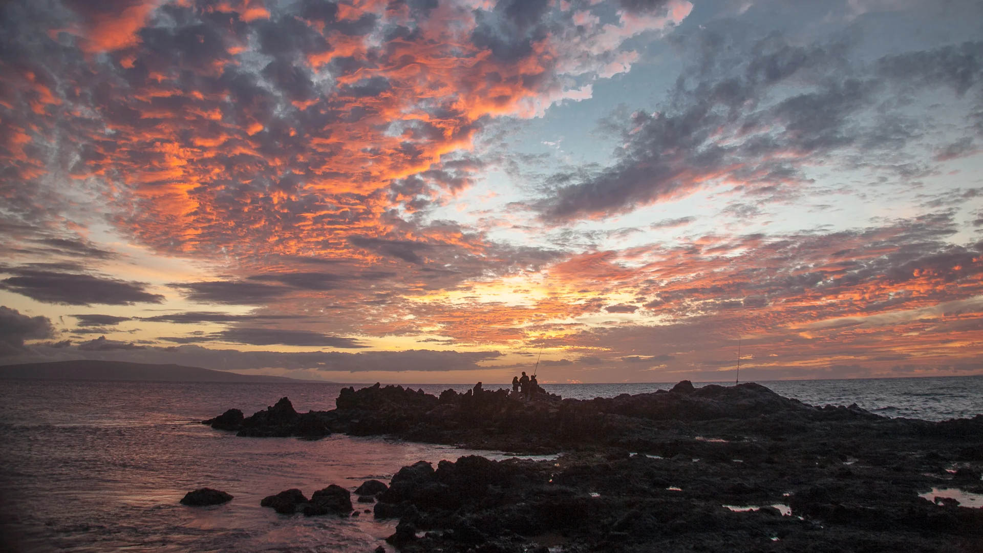 En flammende solnedgang over havet i South Maui, silhuetter af lavaklipper og to lystfiskere ved vandkanten nær Kihei