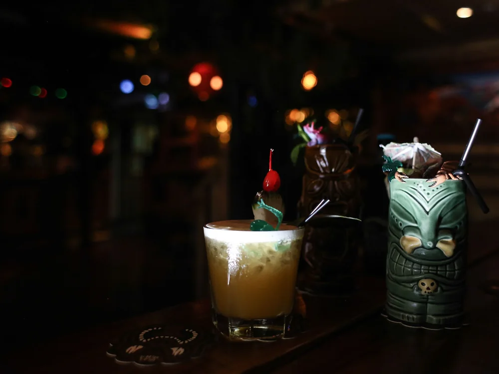 En tropisk cocktail med et kirsebær som garniture på en barbord ved siden af udskårne tiki-krus